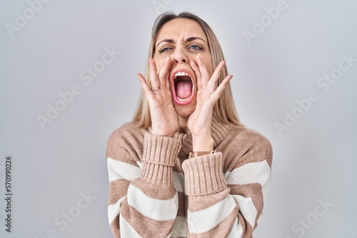 Young blonde woman wearing turtleneck sweater over isolated background shouting angry out loud with hands over mouth