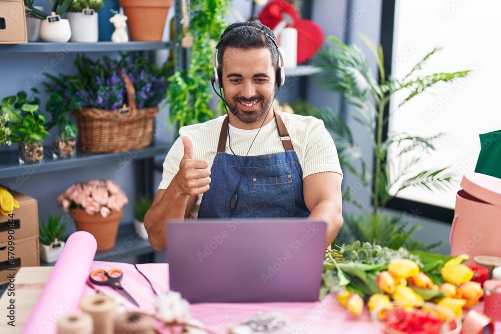 Hispanic man with beard working at florist shop using laptop smiling happy and positive, thumb up doing excellent and approval sign