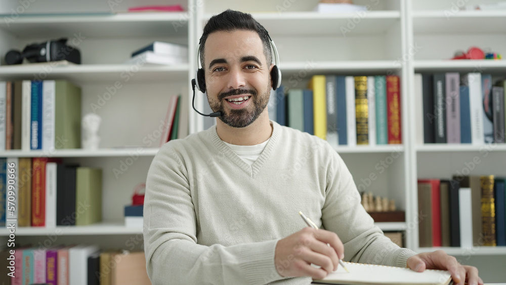 Naklejka premium Young hispanic man student having video call writing on notebook at library university