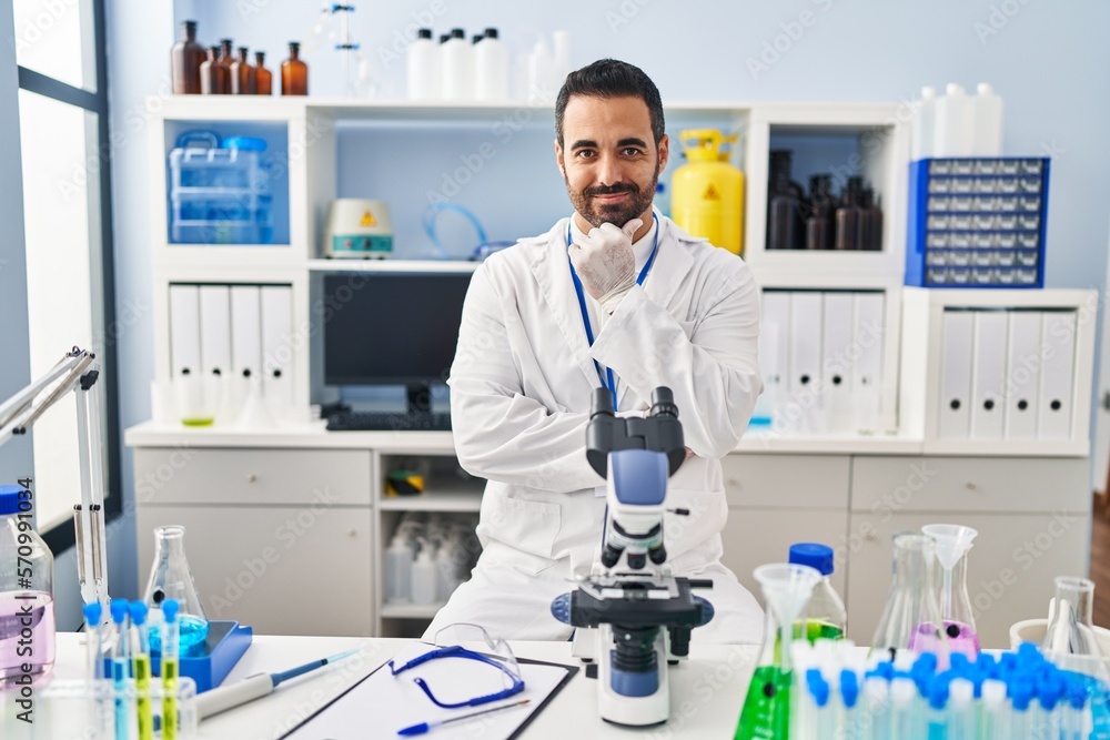 Young hispanic man with beard working at scientist laboratory looking confident at the camera with smile with crossed arms and hand raised on chin. thinking positive. Young hispanic man with beard working at scientist laboratory looking confident at the camera with smile with crossed arms and hand raised on chin. thinking positive.