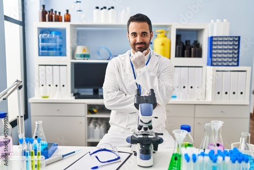 Young hispanic man with beard working at scientist laboratory looking confident at the camera with smile with crossed arms and hand raised on chin. thinking positive.