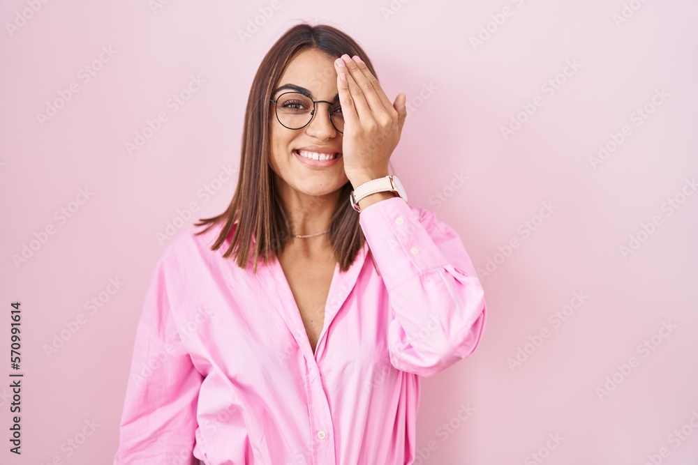 Young hispanic woman wearing glasses standing over pink background covering one eye with hand, confident smile on face and surprise emotion.