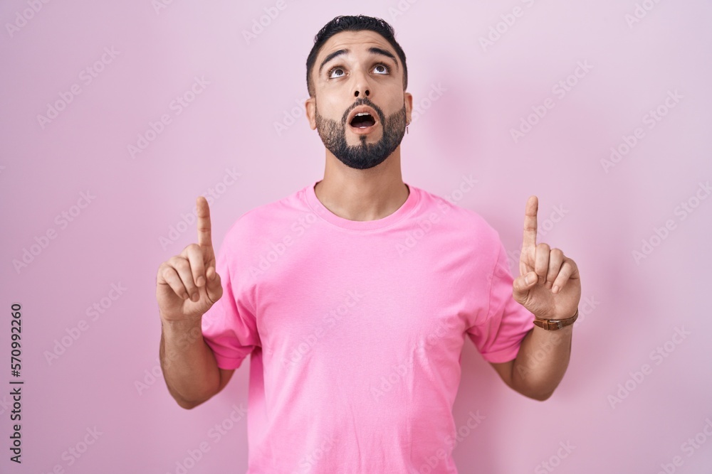 Hispanic young man standing over pink background amazed and surprised looking up and pointing with fingers and raised arms.