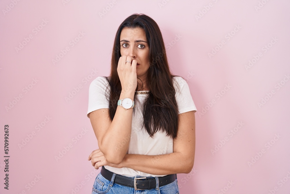 Young brunette woman standing over pink background looking stressed and nervous with hands on mouth biting nails. anxiety problem. Young brunette woman standing over pink background looking stressed and nervous with hands on mouth biting nails. anxiety problem.