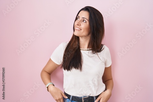 Young brunette woman standing over pink background smiling looking to the side and staring away thinking.