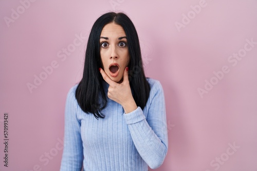 Hispanic woman standing over pink background looking fascinated with disbelief  surprise and amazed expression with hands on chin