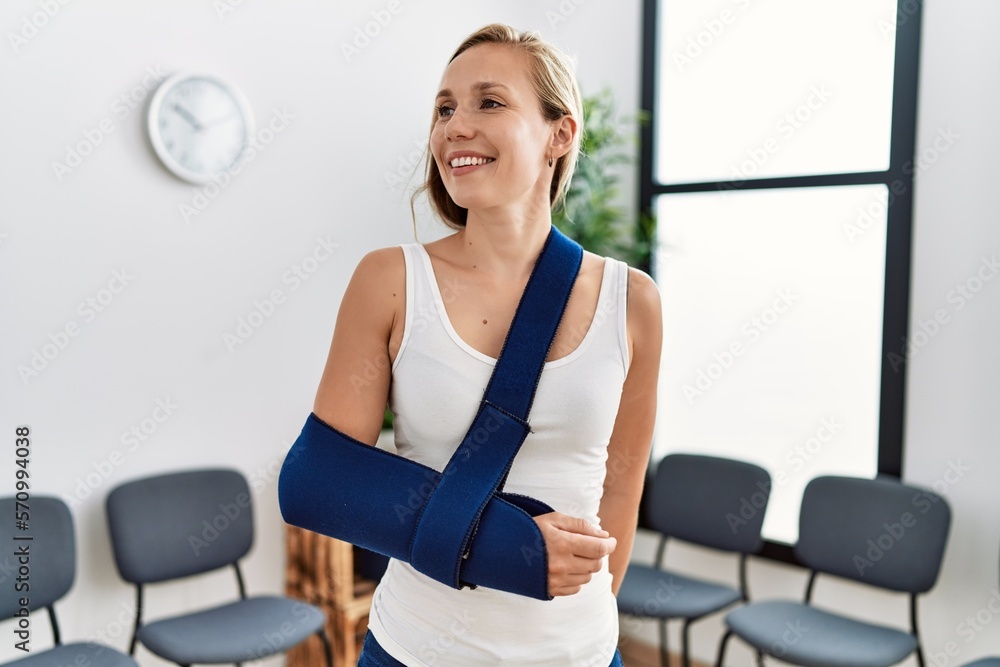 Young caucasian woman wearing sling on arm for accident standing at clinic waiting room