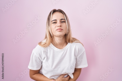 Young blonde woman standing over pink background with hand on stomach because indigestion, painful illness feeling unwell. ache concept.