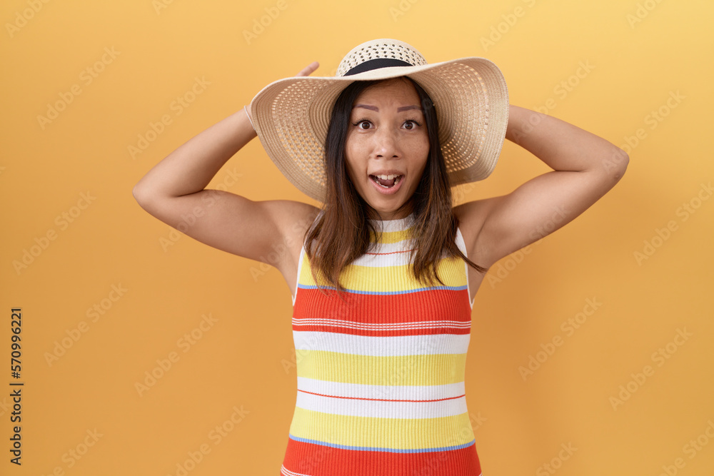 Middle age chinese woman wearing summer hat over yellow background crazy and scared with hands on head, afraid and surprised of shock with open mouth Middle age chinese woman wearing summer hat over yellow background crazy and scared with hands on head, afraid and surprised of shock with open mouth