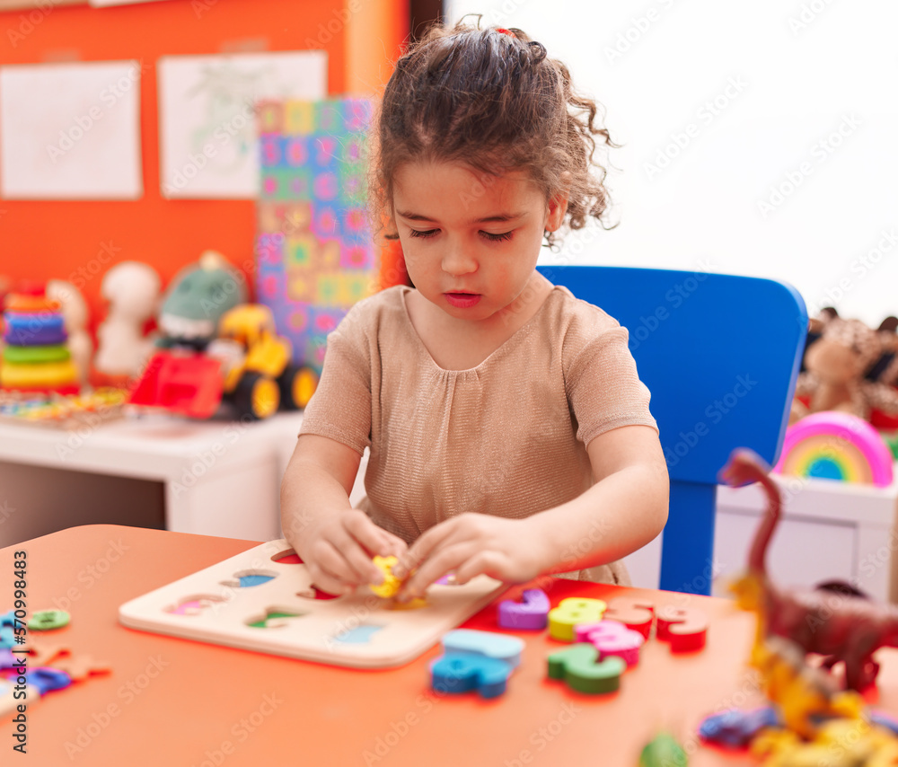 Fototapeta premium Adorable hispanic girl playing with maths puzzle game sitting on table at kindergarten
