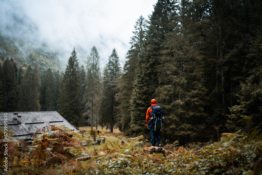 A hiker with orange raincoat is exploring the surroundings of an isolated mountain hut in the forest of Val di Genova, during a rainy and foggy autumnal day, Trentino Alto Adige, Northern Italy