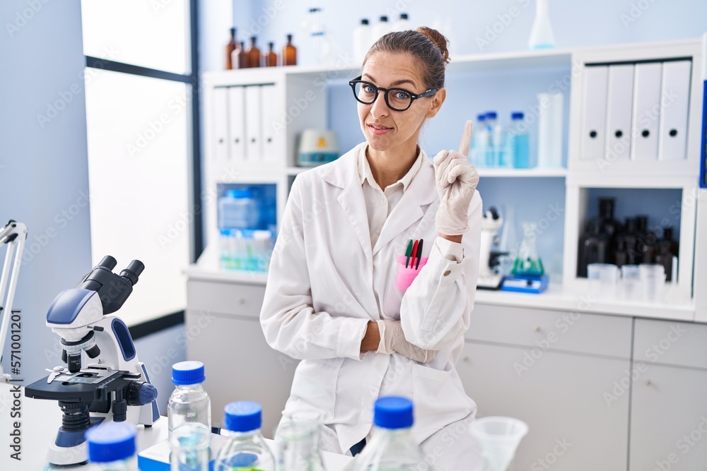 Young woman working at scientist laboratory smiling with an idea or question pointing finger up with happy face, number one Young woman working at scientist laboratory smiling with an idea or question pointing finger up with happy face, number one