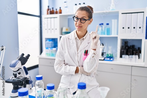 Young woman working at scientist laboratory smiling with an idea or question pointing finger up with happy face  number one