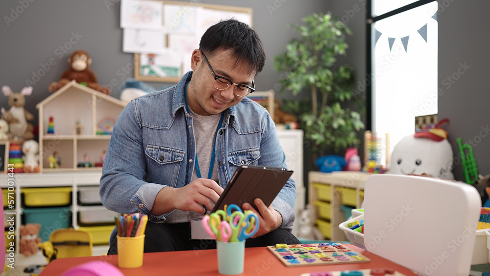 Young chinese man preschool teacher using touchpad sitting on table at kindergarten Young chinese man preschool teacher using touchpad sitting on table at kindergarten