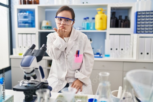 Hispanic girl with down syndrome working at scientist laboratory looking at the camera blowing a kiss with hand on air being lovely and sexy. love expression.
