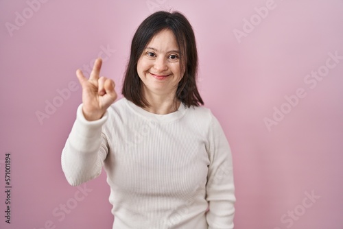 Woman with down syndrome standing over pink background showing and pointing up with fingers number two while smiling confident and happy.
