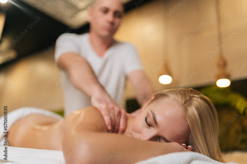 Obraz premium Low-angle view of naked blonde female getting professional relaxing massage at spa salon lying on massage table with closed eyes. Male masseur performing circular movements to relax back muscles.