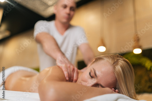 Low-angle view of naked blonde female getting professional relaxing massage at spa salon lying on massage table with closed eyes. Male masseur performing circular movements to relax back muscles.