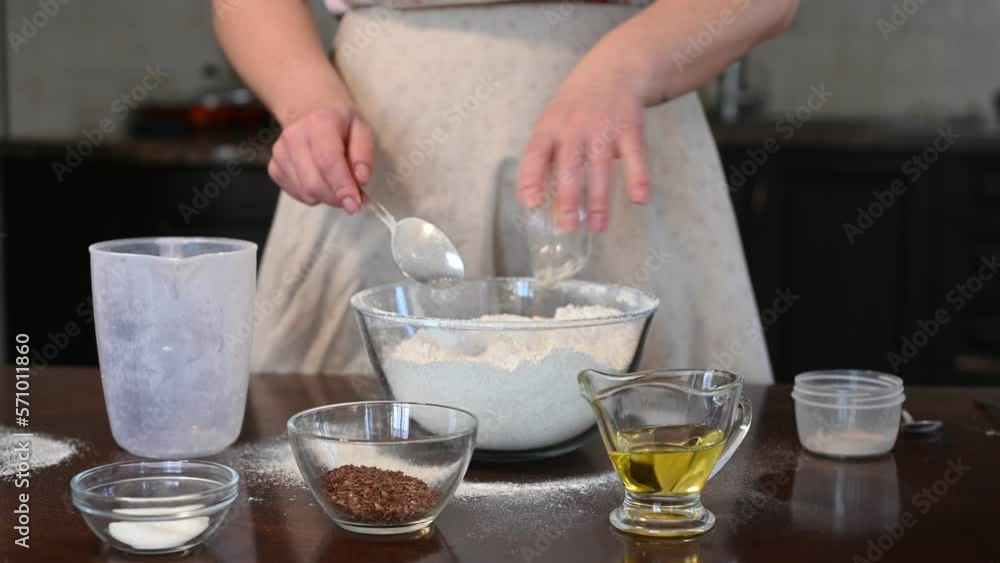 Woman prepares homemade bread with flax seeds. He gradually adds ingredients - salt, sugar, yeast, seeds. Mixes ingredients with flour in a transparent container. Home baking, food, traditions Woman prepares homemade bread with flax seeds. He gradually adds ingredients - salt, sugar, yeast, seeds. Mixes ingredients with flour in a transparent container. Home baking, food, traditions