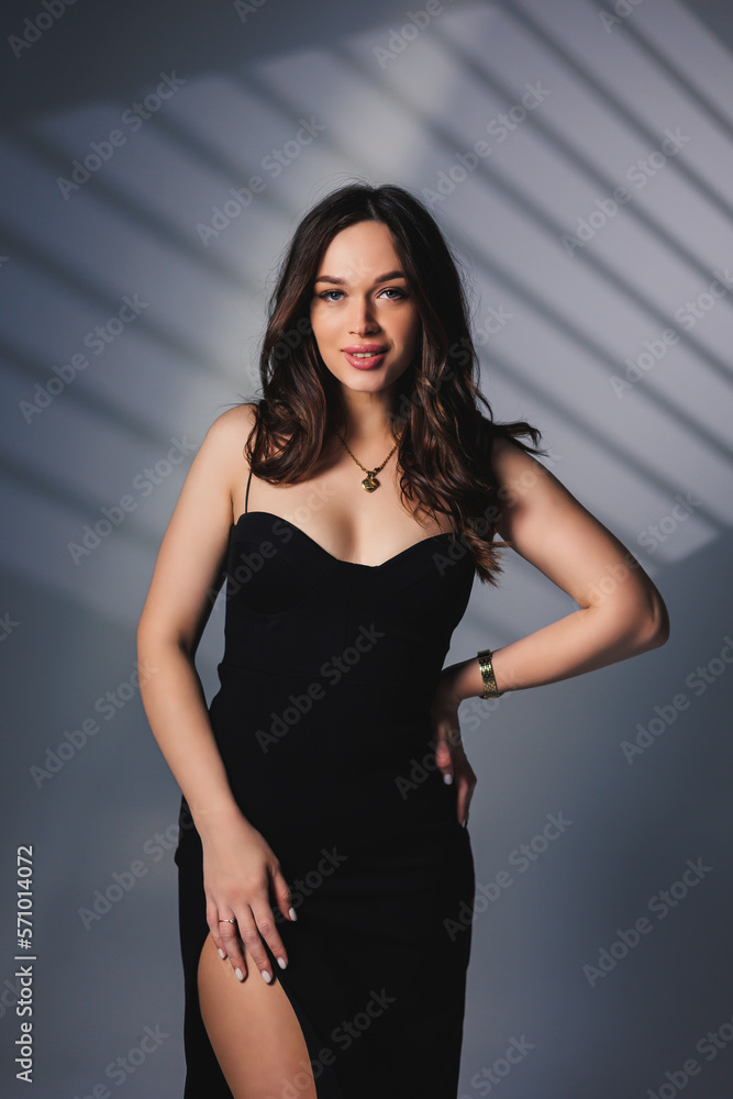 Fototapeta premium A young pretty woman in a black dress with a slit on the leg poses while standing in the studio. Confident attractive woman with makeup and hairstyle