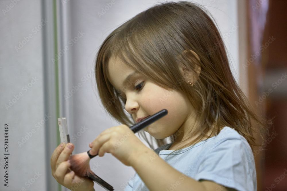 A little girl holds a blush brush to apply powder on her cheeks and face, happily looking in the mirror. Cute baby is playing makeup makeup game.. A little girl holds a blush brush to apply powder on her cheeks and face, happily looking in the mirror. Cute baby is playing makeup makeup game..