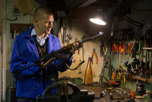 Focused craftsman of weapons workshop inspecting Kalashnikov assault rifle brought to him for repairs