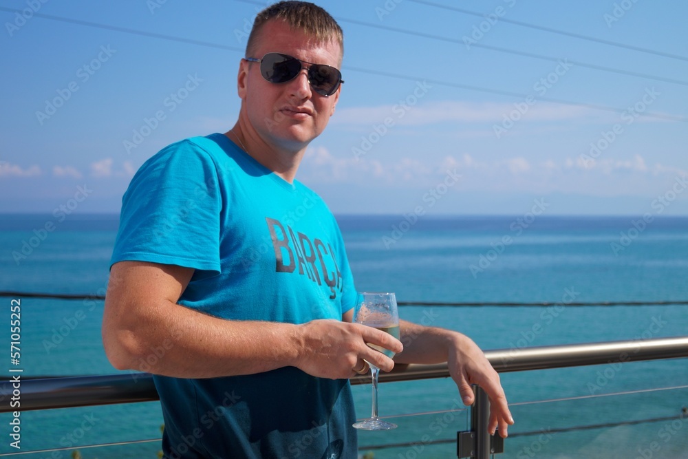 A young man in sunglasses on the balcony. Against the backdrop of the blue sea in Greece. A glass of white wine in hand. Concept of rest.