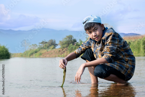 Fototapeta Naklejka Na Ścianę i Meble -  Asian boy in a plaid shirt wearing a cap holds freshwater algae that he plucks from a river in the area of his village to study the river's cleanliness, ecosystem, ecoeffect and pesticide effect.