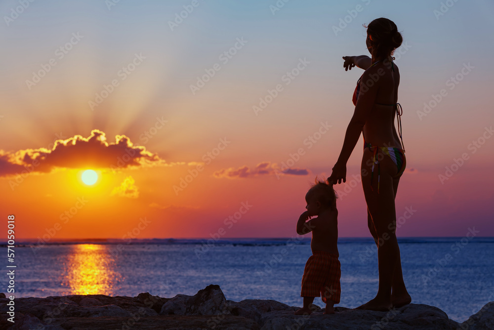 Φωτογραφία Black silhouette of happy family walking on sea beach - mother hold baby son hand, point sun and look at sunset sky