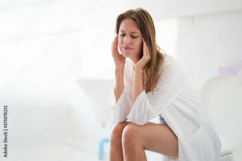 Young Caucasian woman sitting at the bathroom close her eyes while rubbing her head because of migraine. Young Caucasian woman sitting at the bathroom close her eyes while rubbing her head because of migraine.