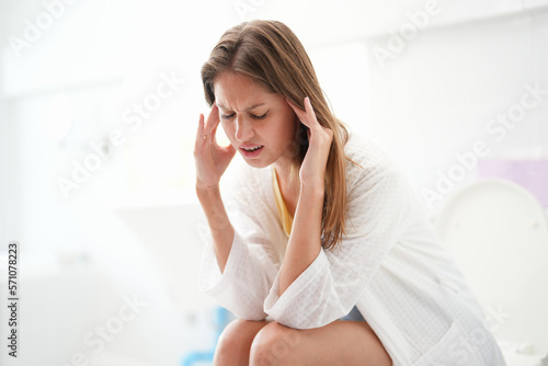 Young Caucasian woman sitting at the bathroom while rubbing her head because of migraine.