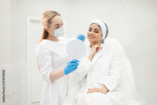 cosmetologist doctor and patient girl look in the mirror at face skin in cosmetology clinic and smile