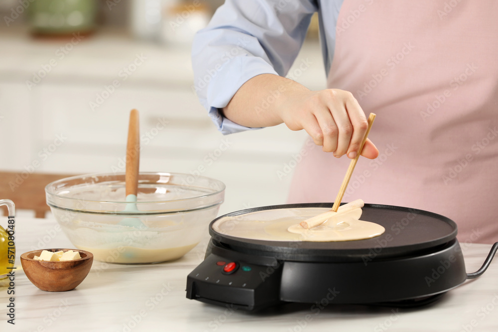 Woman cooking delicious crepe on electric pancake maker at white marble table in kitchen, closeup Woman cooking delicious crepe on electric pancake maker at white marble table in kitchen, closeup