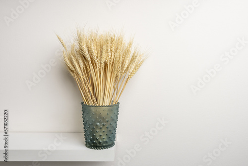 A decorative blue glass container containing a bunch of ears of dried wheat on a white shelf and white wall