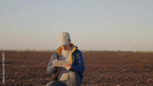Man agronomist squats on black ground plot of plowed and cultivated field in countryside on spring day. Farmer enters data for report on soil condition into tablet. Agricultural business concept