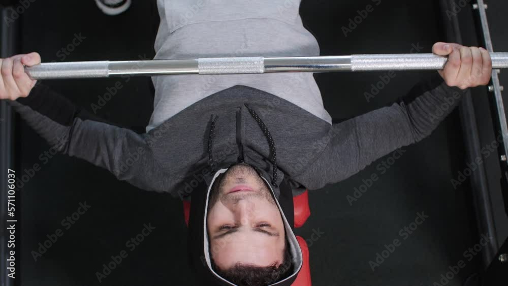 Young sportive strong man in hoodie is doing bench press exercise in gym on his training, top view. Sport workout, healthy lifestyle, strength and power training concept. He lifts the bar with effort. Young sportive strong man in hoodie is doing bench press exercise in gym on his training, top view. Sport workout, healthy lifestyle, strength and power training concept. He lifts the bar with effort.