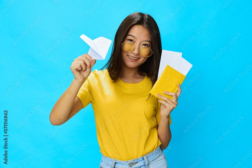 Asian woman tourist holding a paper airplane and passport with plane ticket in hand with yellow sunglasses smile with teeth in yellow T-shirt and blue jeans on blue background, copy space Asian woman tourist holding a paper airplane and passport with plane ticket in hand with yellow sunglasses smile with teeth in yellow T-shirt and blue jeans on blue background, copy space