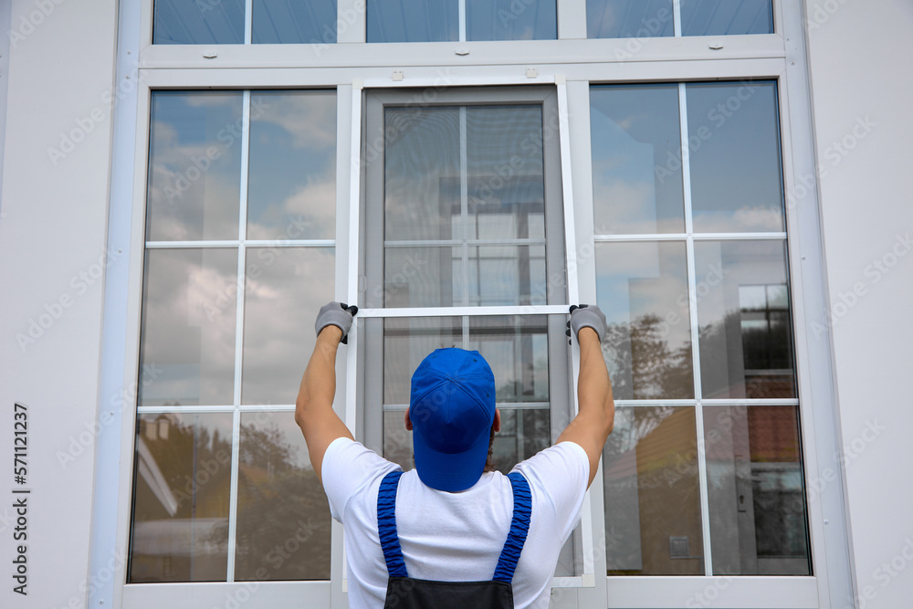 Obraz na plátně Background of blue-cap worker installing a mosquito net on large plastic window outside the building in summer