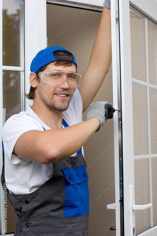 Caucasian handsome male worker stands at open door in overalls and protective glasses screws fittings with screwdriver. Experienced foreman performs work of servicing the locks on the plastic door Caucasian handsome male worker stands at open door in overalls and protective glasses screws fittings with screwdriver. Experienced foreman performs work of servicing the locks on the plastic door