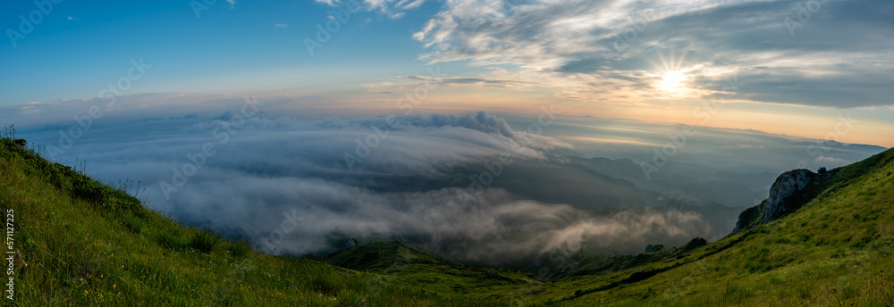 Panorama of green meadows with rocks and rocky mountains with fog in the valley and clouds in the sky in romanian mountains in muntii ciucas