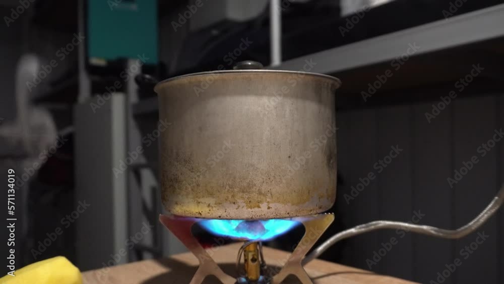 A man's hand lights a gas burner from a match. There is an old pot on the burner. The hose is connected to the gas cylinder. Cooking food in an escape capsule. There are potatoes. Blue flame of fire A man's hand lights a gas burner from a match. There is an old pot on the burner. The hose is connected to the gas cylinder. Cooking food in an escape capsule. There are potatoes. Blue flame of fire