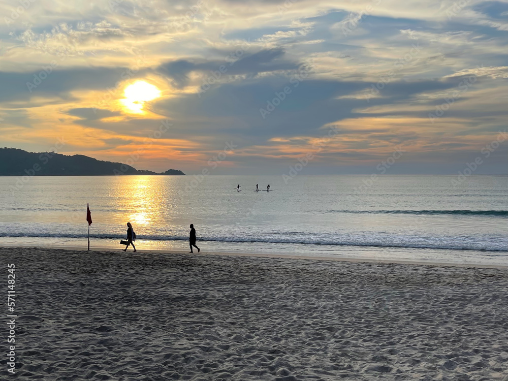 Beautiful sunset. Tropical beach. People walk along the sea. Three men are swimming with SUP (paddle boards) on the water surface in the distance. Bright sun glow and amazing clouds on the sky. 