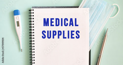 Notepad, medical mask, thermometer and pen on a blue background. MEDICAL SUPPLIES text in a notebook.