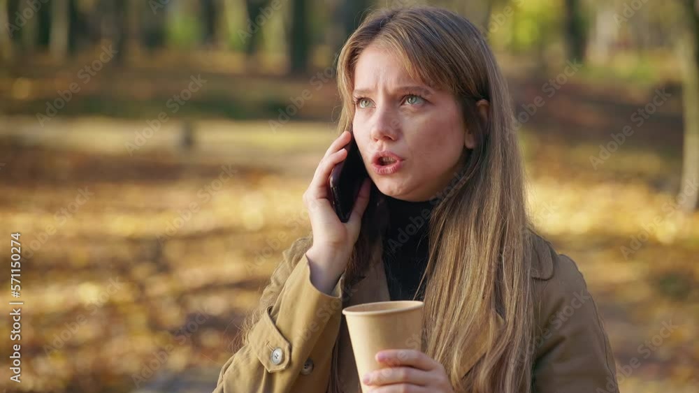 Side view of angry, young lady talking on smartphone in park. Attractive girl standing, using smartphone, drinking coffee, holding paper cup. Concept of modern lifestyle. Side view of angry, young lady talking on smartphone in park. Attractive girl standing, using smartphone, drinking coffee, holding paper cup. Concept of modern lifestyle.