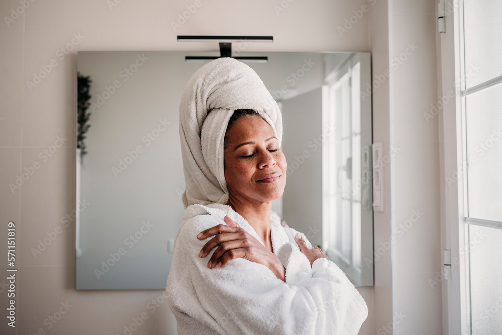 Smiling mature woman with eyes closed hugging self in bathroom at home Smiling mature woman with eyes closed hugging self in bathroom at home