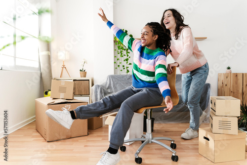 Carefree young woman pushing friend sitting in swivel chair at new home photo