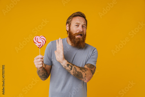 Bearded man doing stop gesture while holding lollipop over background