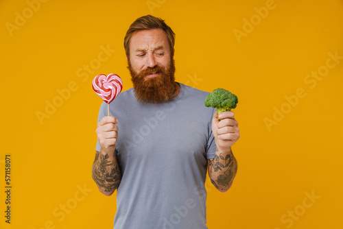 Bearded man holding lollipop and broccoli standing over background