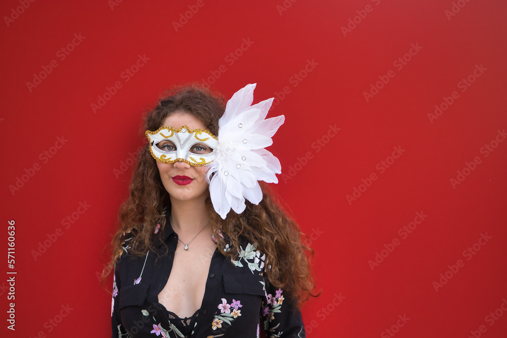 Portrait of a young and beautiful woman, blonde, with curly hair and blue eyes, dressed in flowers and black top, with a white carnival mask with feathers, on a red background.