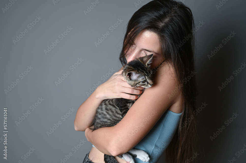 Woman hugging her kitten. Striped kitten lies on on the shoulder of a woman on gray background. Woman hugging her kitten. Striped kitten lies on on the shoulder of a woman on gray background.